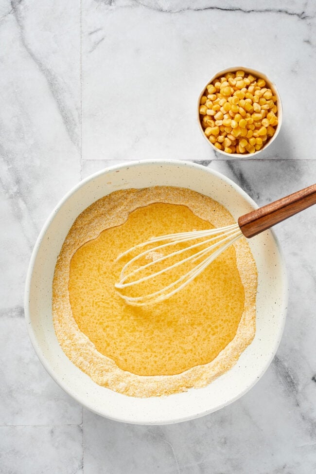 A bowl of the dry ingredients for cornbread, with the wet ingredients poured on top, with a whisk in the bowl, next to a bowl of corn