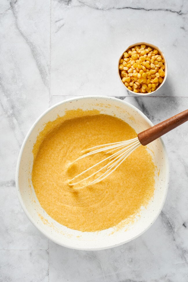 A mixing bowl filled with cornbread batter, without the corn, with a whisk in it, next to a bowl of corn kernels