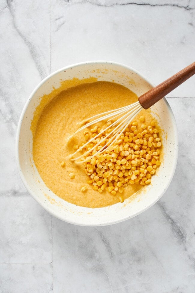 A mixing bowl with cornbread batter, with corn kernels on top, and a whisk