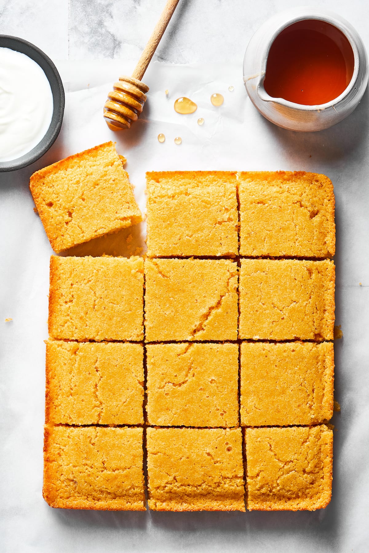 Cornbread cut into 12 pieces, with one corner askew, next to a jar of honey and a honey stirrer