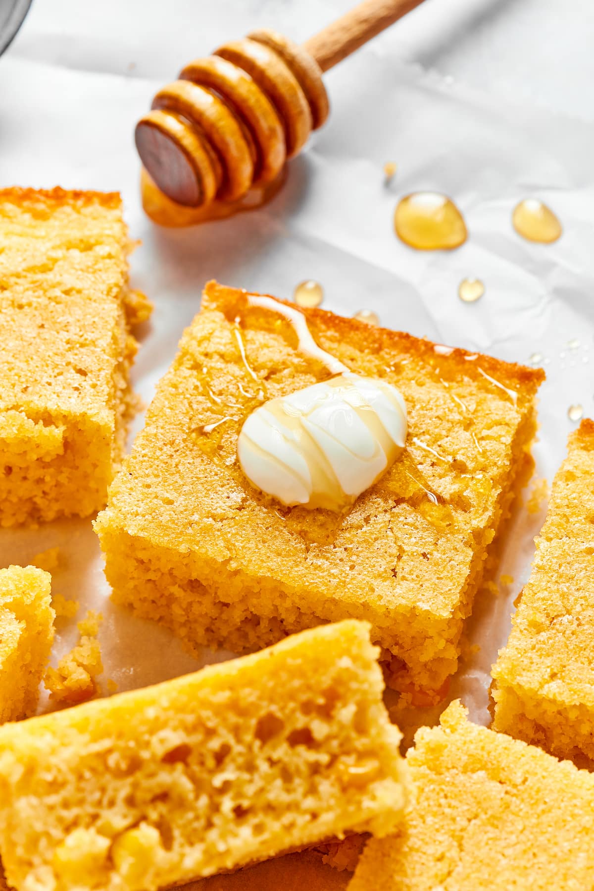 Close up of a square of cornbread, topped with a dollop of creme fraiche and a drizzle of honey, next to other slices and a honey stirrer