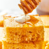 A stack of easy cornbread squares, with a dollop of creme fraiche on top, and a honey stirrer drizzling honey over the top