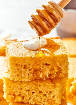 A stack of easy cornbread squares, with a dollop of creme fraiche on top, and a honey stirrer drizzling honey over the top