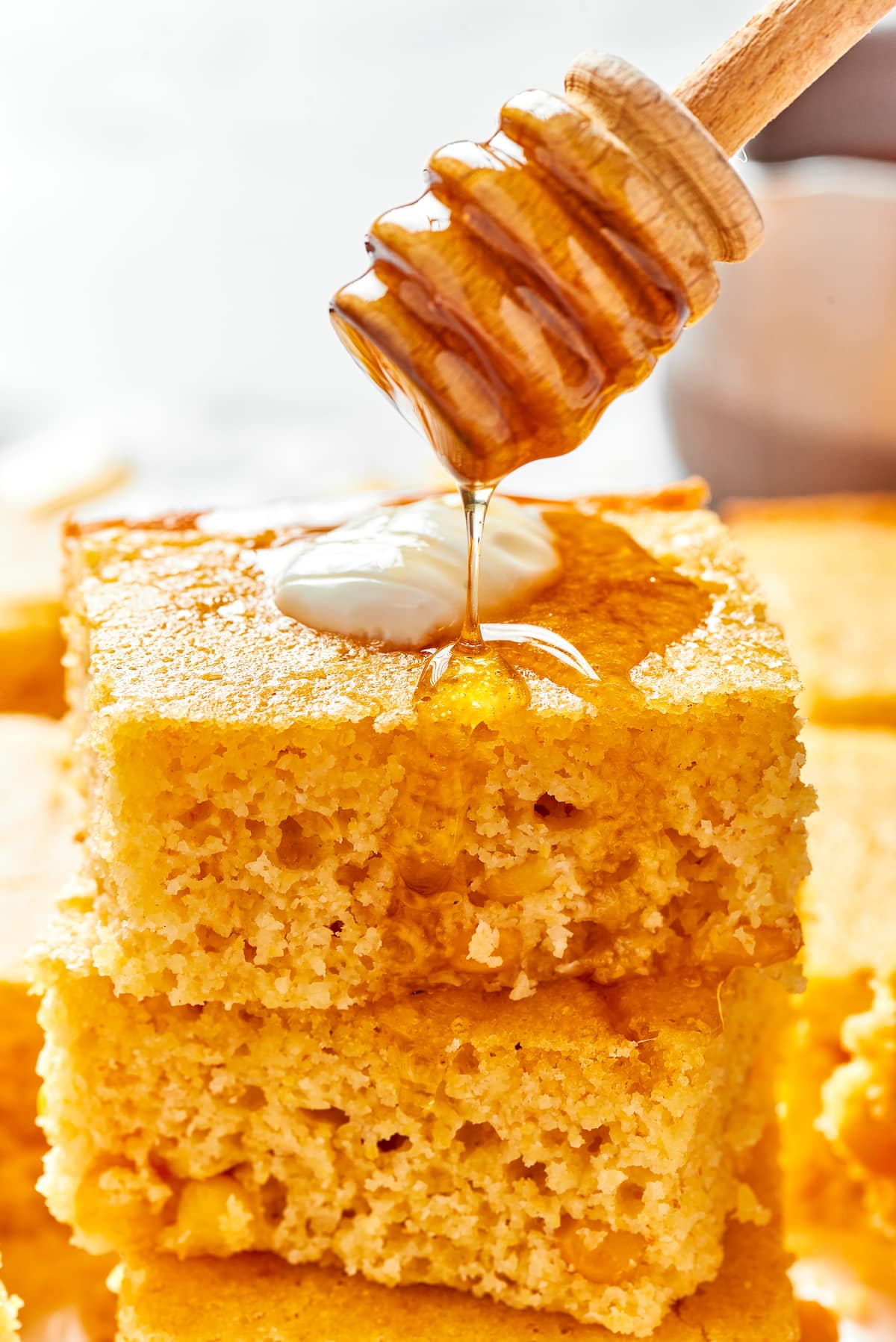 A stack of cornbread squares, with a dollop of creme fraiche on top, and a honey stirrer drizzling honey over the top