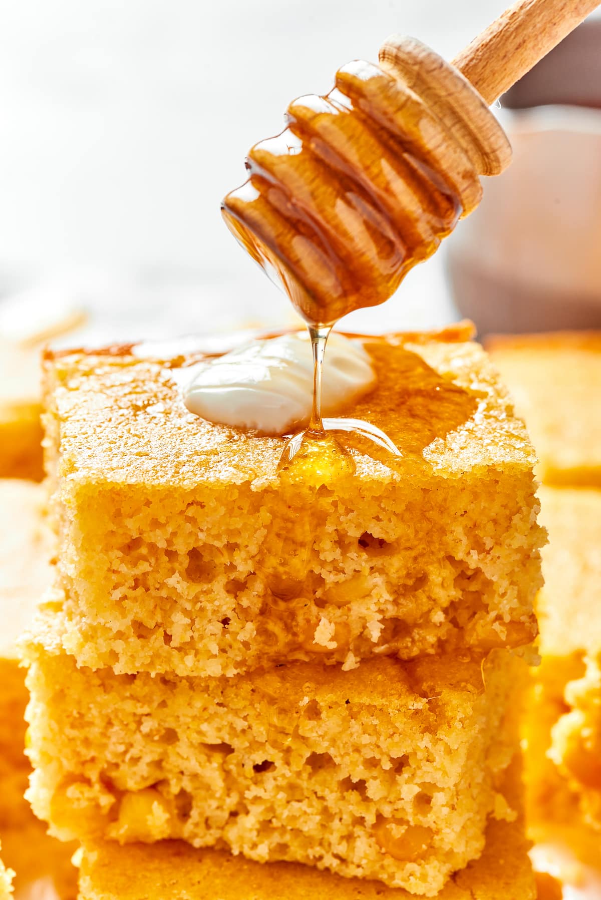 This easy cornbread recipe is tender, moist, perfectly sweet, and takes only 35 minutes to make, making it a wonderful side dish A stack of cornbread squares, with a dollop of creme fraiche on top, and a honey stirrer drizzling honey over the top