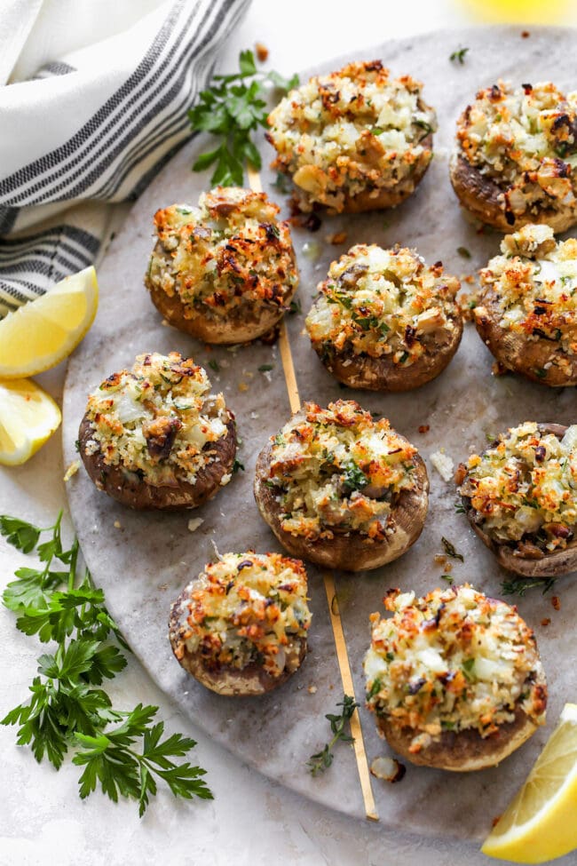 stuffed mushrooms on marble tray with fresh herbs and lemon wedges.