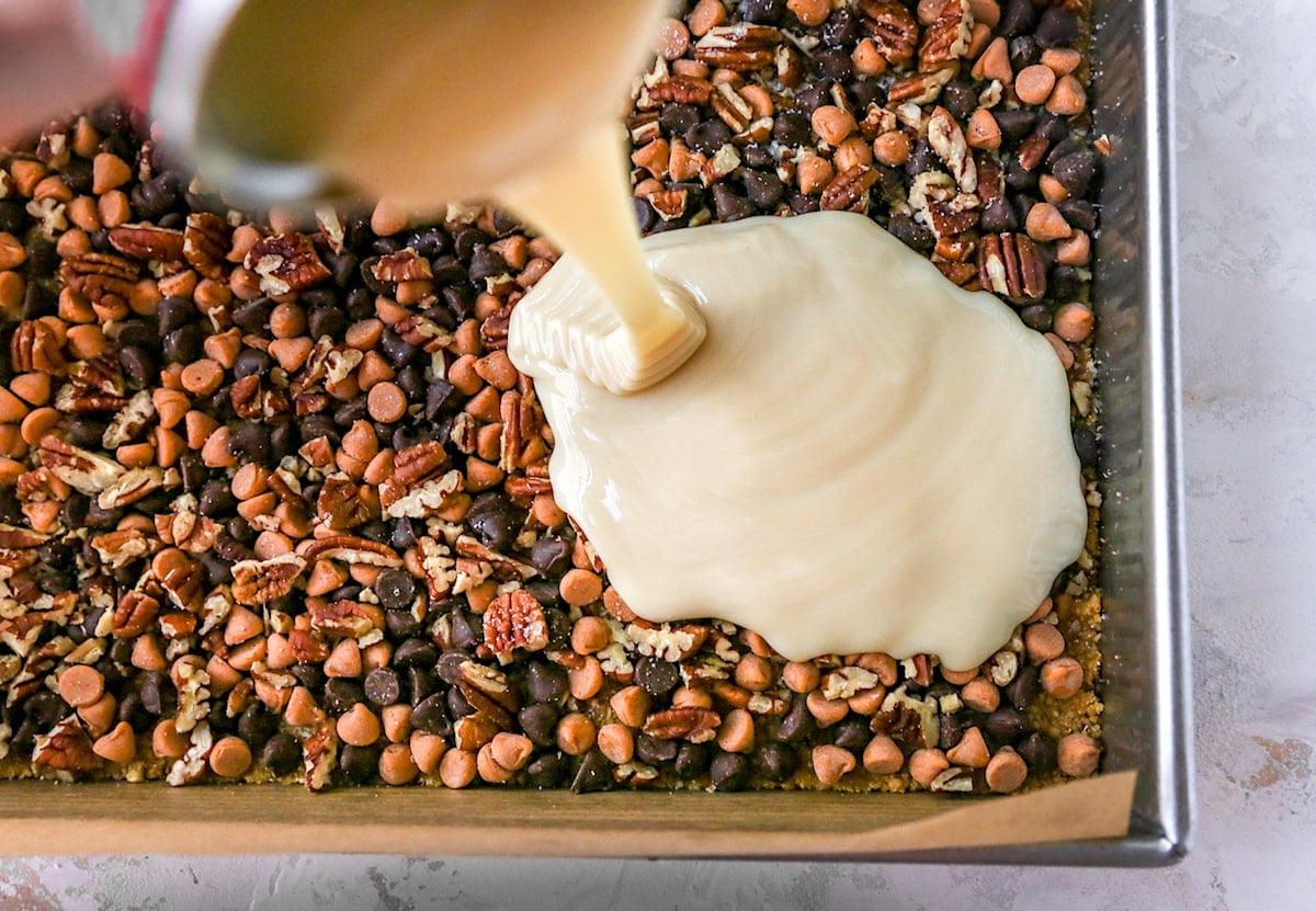 magic cookie bars in pan with sweetened condensed milk being poured over the top. 