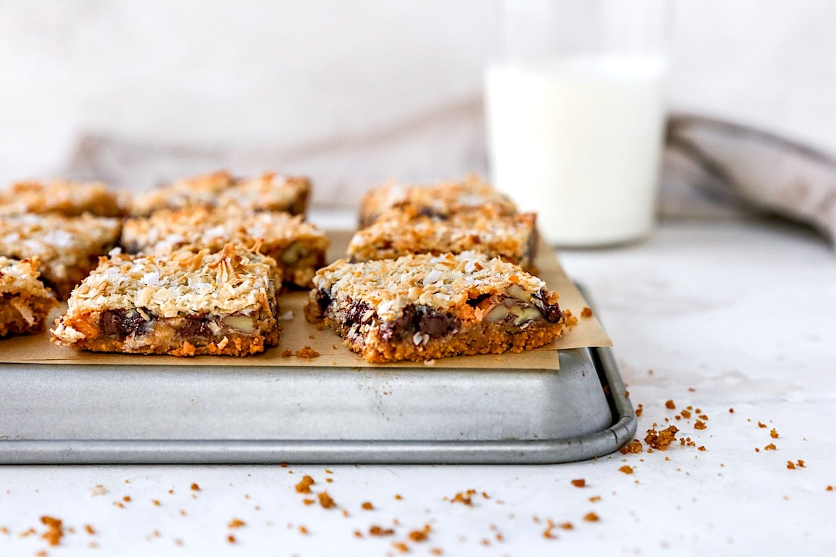 Magic cookie bars cut into squares on baking pan with parchment paper and glass of milk. 