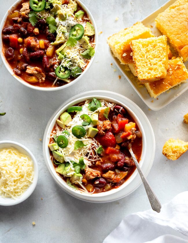 Turkey chili in a bowl with toppings and cornbread on a plate.