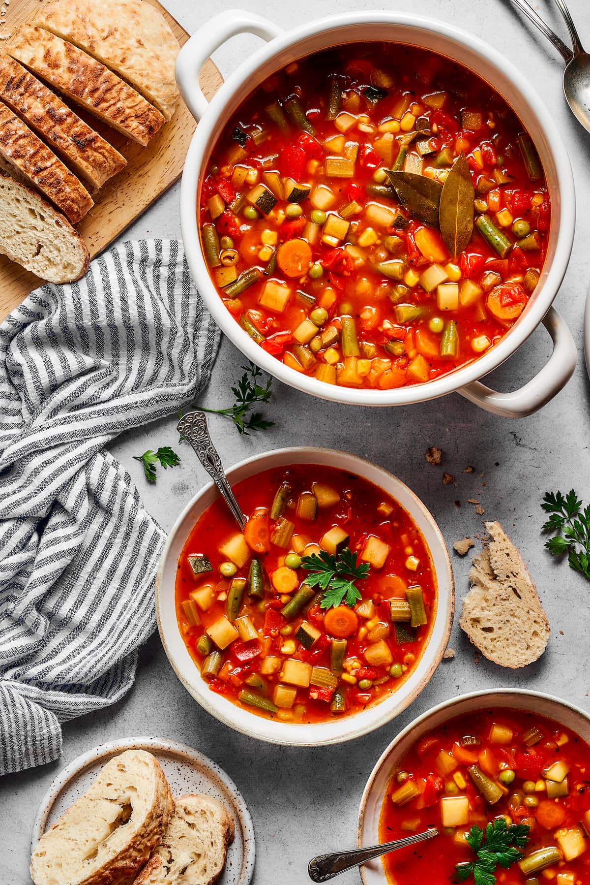 A bowl of homemade vegetable soup with a spoon, next to a large pot. A full spread of homemade vegetable soup in two bowls, next to a large pot and a loaf of crusty bread.