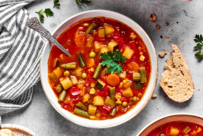 A bowl of homemade vegetable soup with a spoon, next to a bread slice.