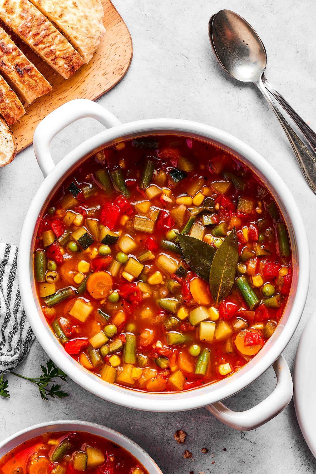 A pot of vegetable soup next to a loaf of crusty bread and a spoon.