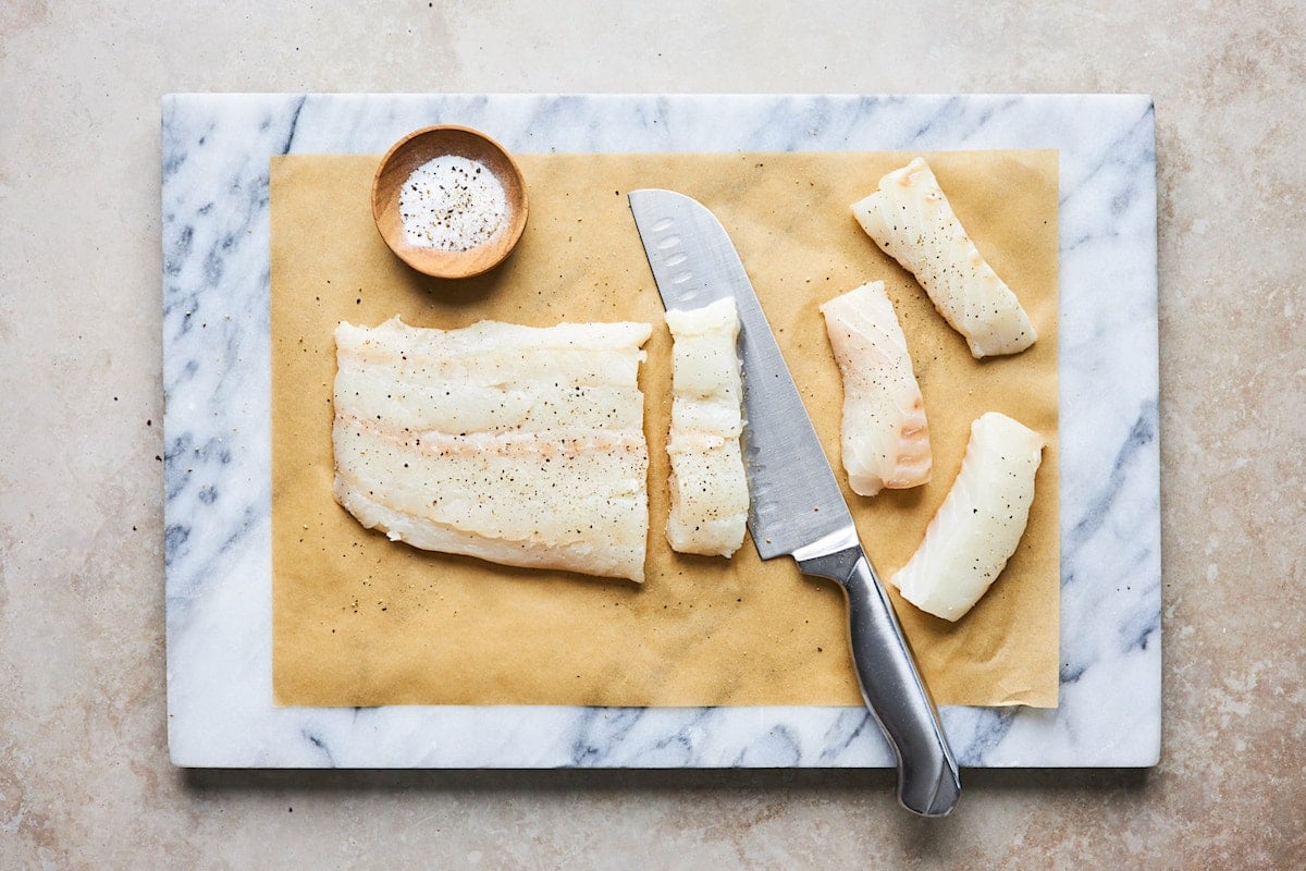 white fish on cutting board being cut into sticks with sharp knife. 