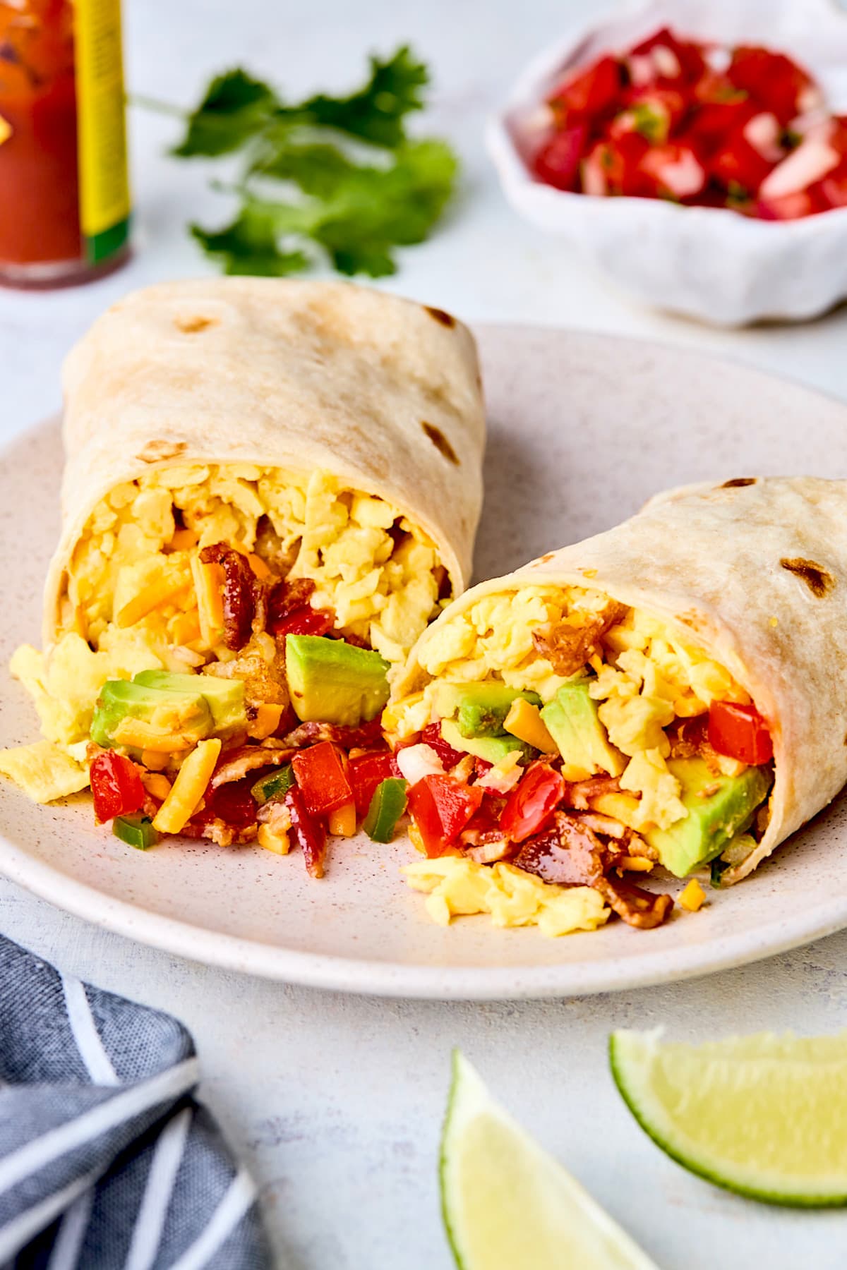 breakfast burrito cut in half with eggs, bacon, tater tots, cheese, avocado, and pico de gallo on plate. 