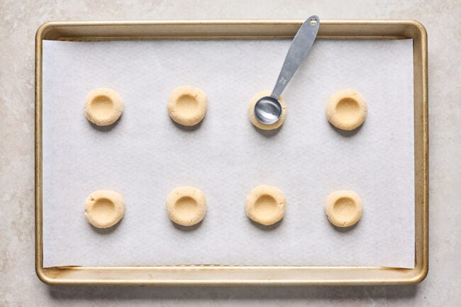 thumbprint cookie dough pressed into thumbprints with a teaspoon on a baking sheet lined with parchment paper. 