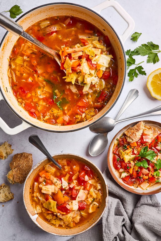 cabbage soup in pot with soup in two bowls with crusty bread and fresh herbs.