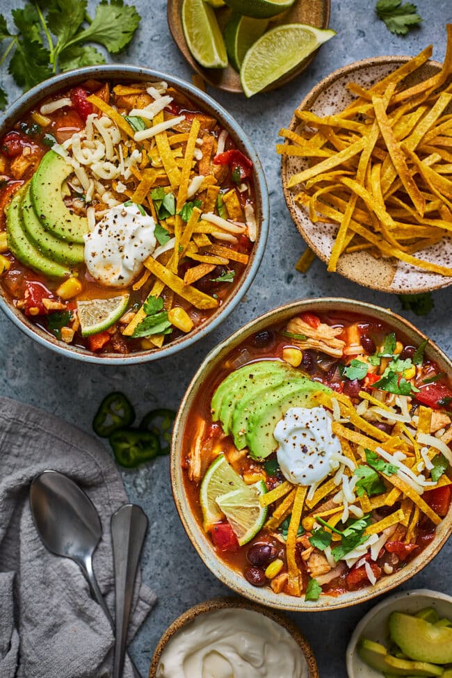 chicken tortilla soup in two bowls with tortilla strips, sour cream, cheese, cilantro, lime and avocado. 