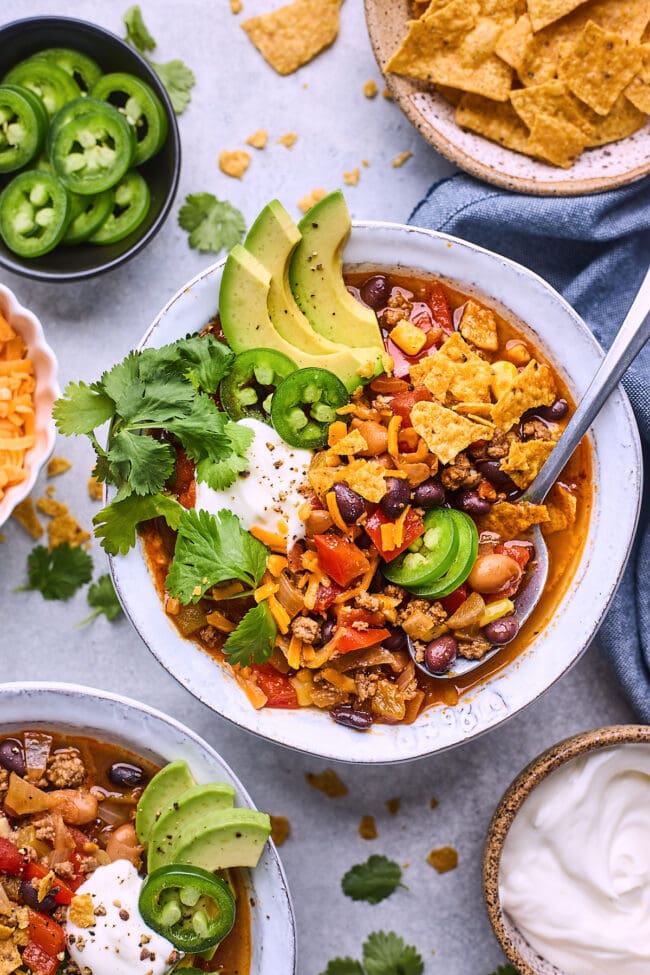 taco soup in a bowl with a spoon and sour cream, avocado slices, chips, cheese, cilantro, and jalapeño slices. 