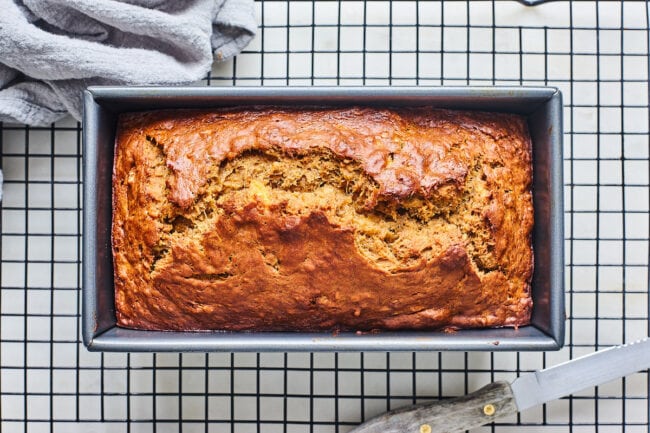 banana bread in pan on cooling rack with bread knife and tea towel.