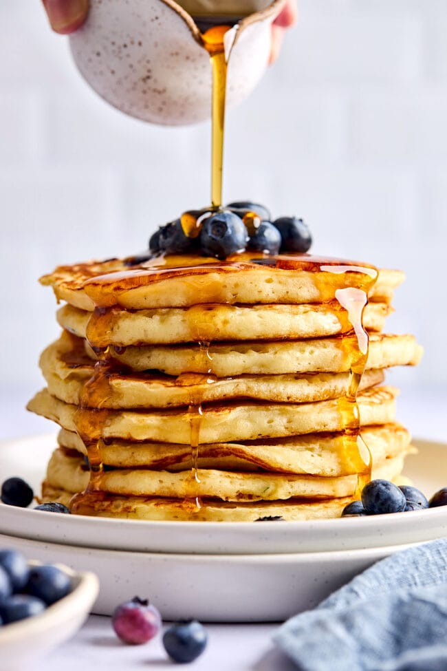 stack of blueberry pancakes on plate with maple syrup being poured over the top.
