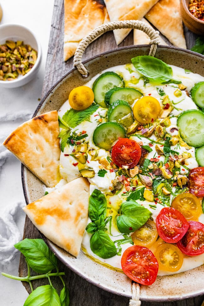 whipped feta in bowl with tomatoes, cucumbers, herbs, and pita bread.