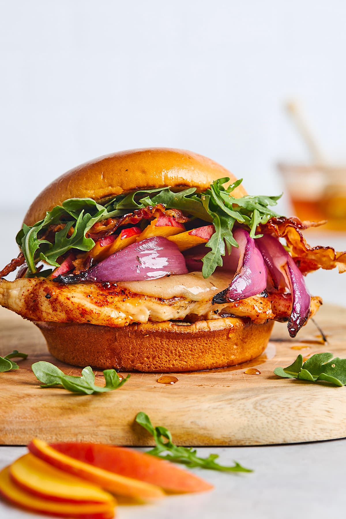 close up of grilled chicken sandwich on bun with cheese, arugula, red onions, bacon, and peaches.
