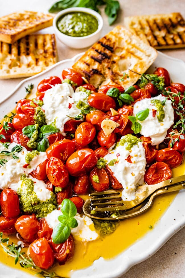 roasted tomatoes with burrata and pesto on platter with fork and spoon.