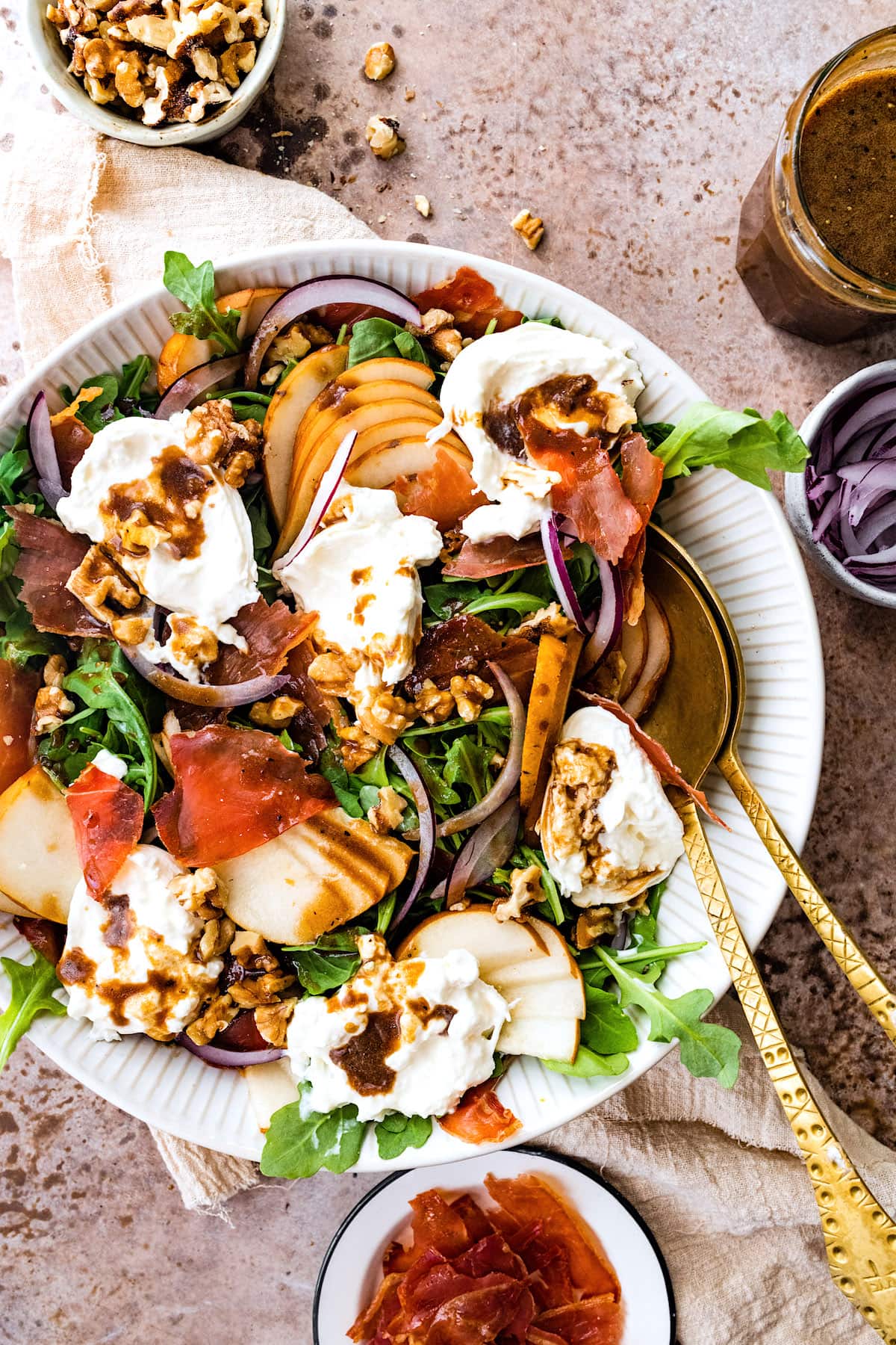 crispy prosciutto salad with pears, burrata, red onion, and walnuts in a bowl with balsamic vinaigrette and gold serving spoons. 