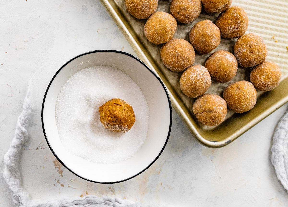 pumpkin gingersnap cookie dough balls on a baking sheet and one in a bowl of sugar. 