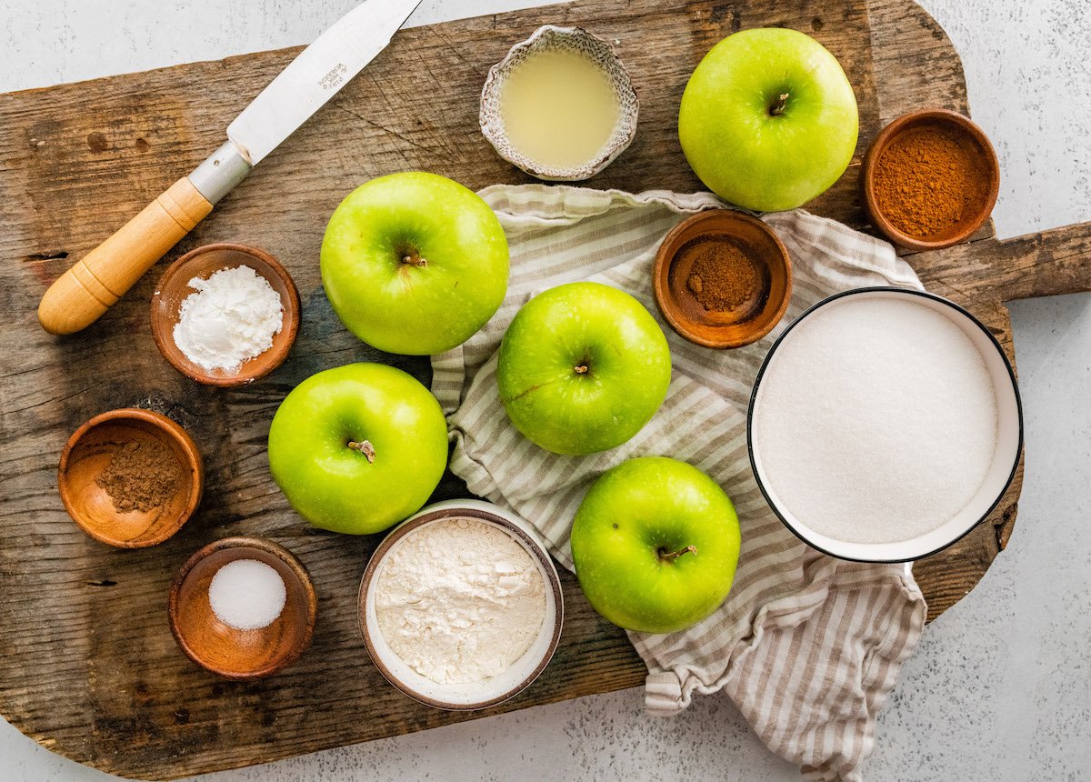 apples, spices, flour, and sugar in bowls on wood cutting board with a knife and kitchen towel. 