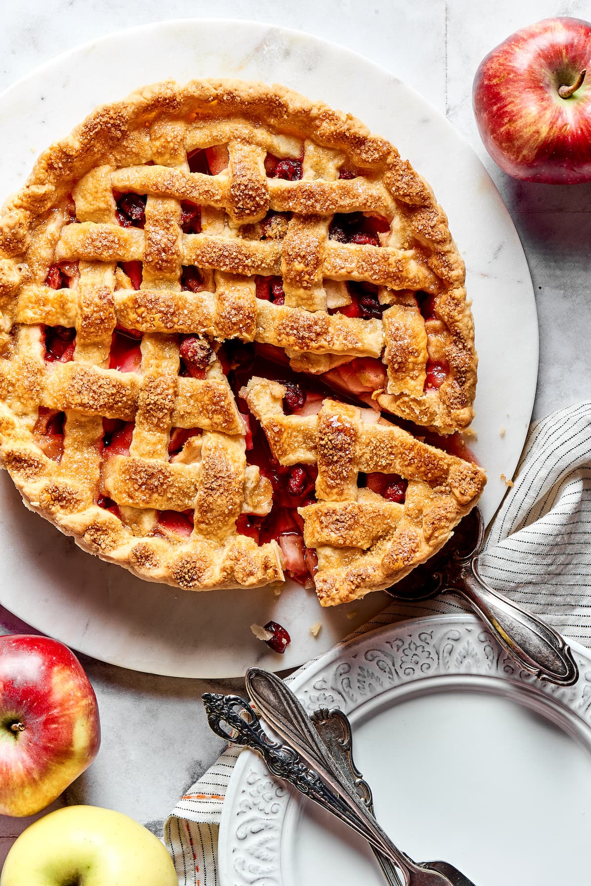 apple cranberry pie with a piece cut out on marble board and plate and forks on the side.