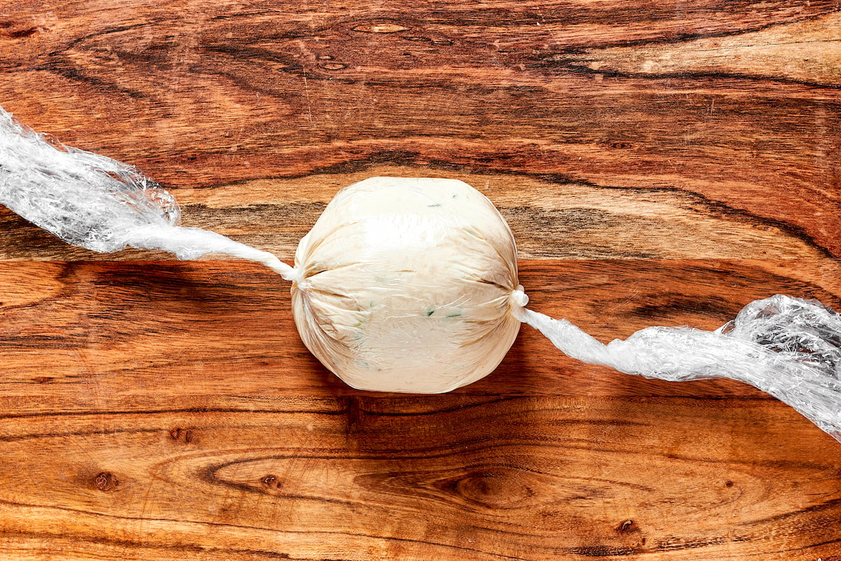 cheese ball being formed in plastic wrap on wood cutting board. 