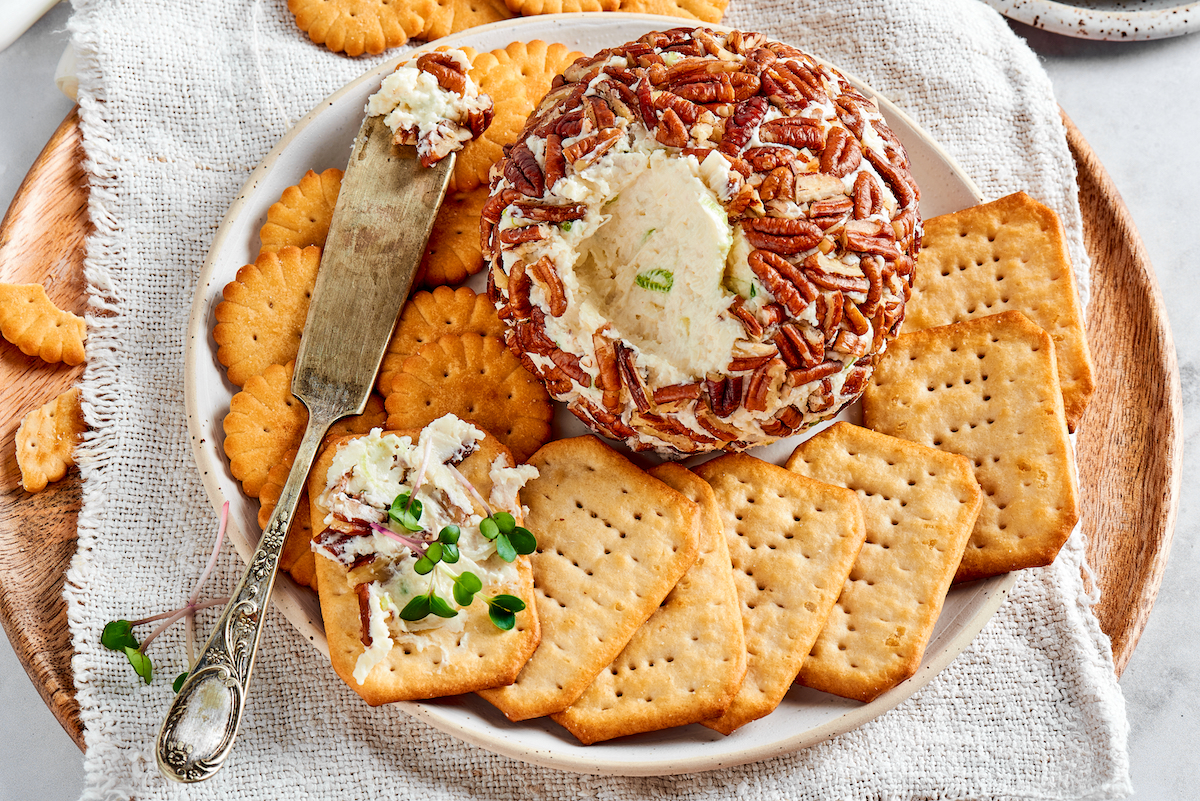 cheese ball covered in pecans on plate with cheese spread on a cracker. 