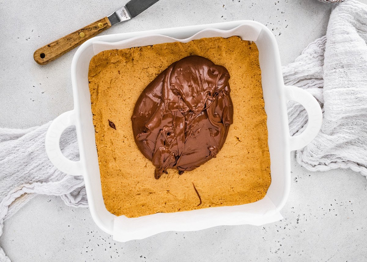 no bake peanut butter bars in pan with melted chocolate in the center getting ready to be spread with spatula.