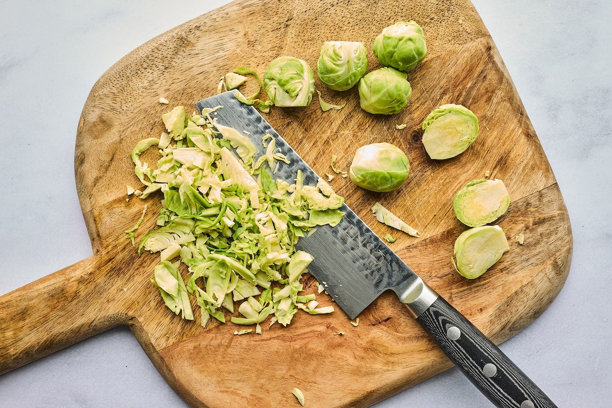 Brussels sprouts on wood cutting board with large knife being shredded. 