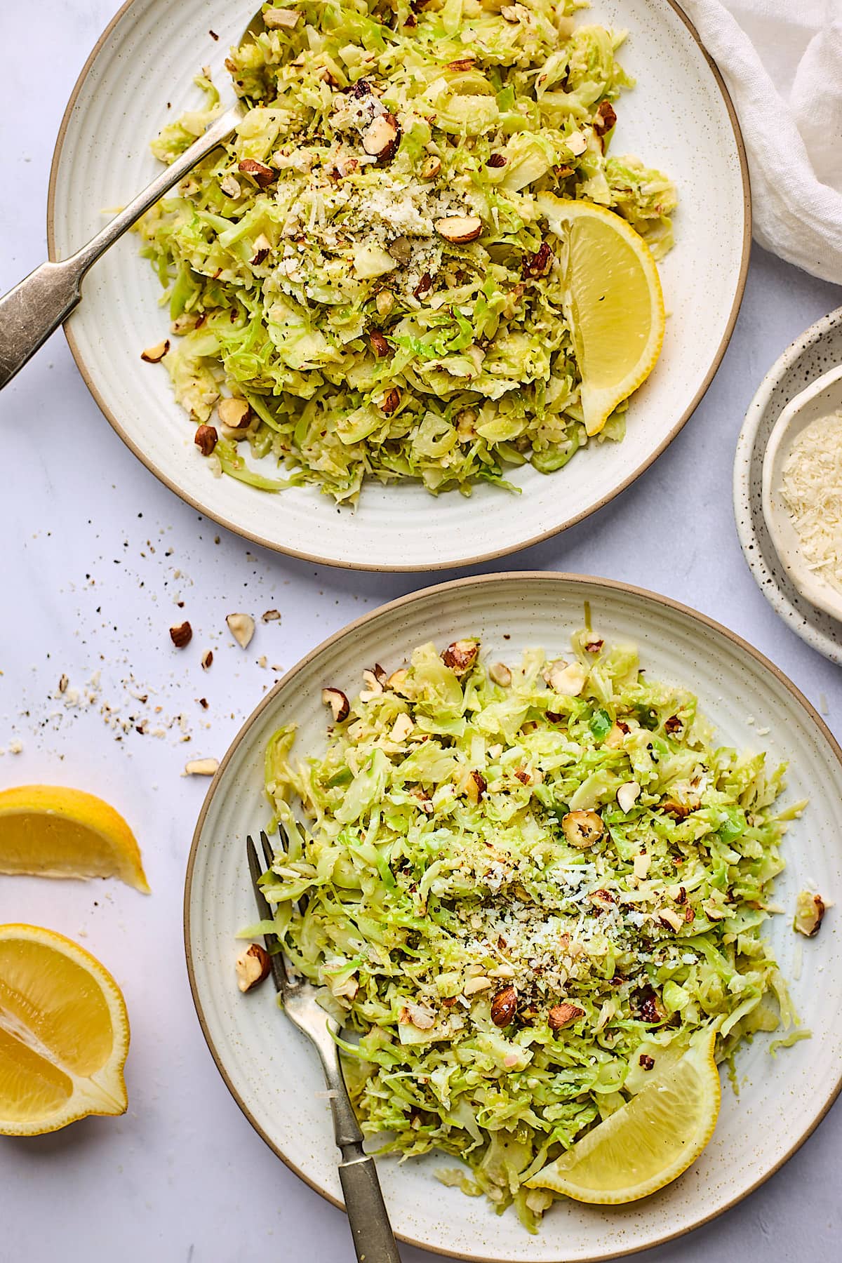sautéed shredded Brussels sprouts with Parmesan cheese and hazelnuts on plates with forks and lemon wedges.
