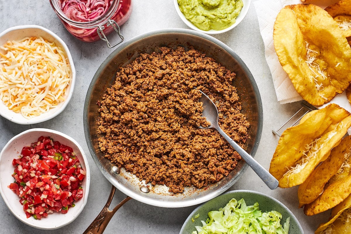 ground beef taco meat in skillet with spoon and toppings in bowls with crispy taco shells.