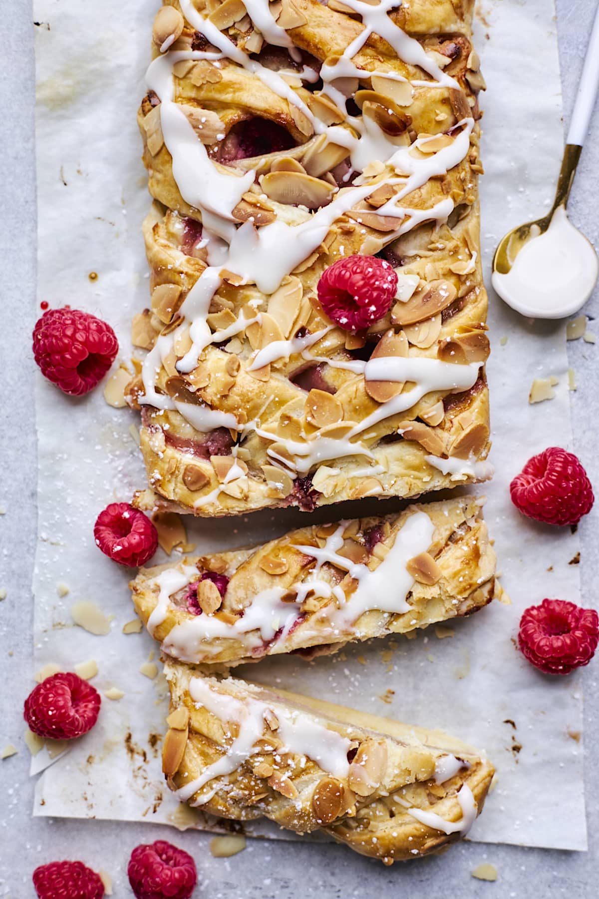 raspberry cream cheese breakfast braid on parchment paper with sliced almonds, almond glaze, and fresh raspberries. 