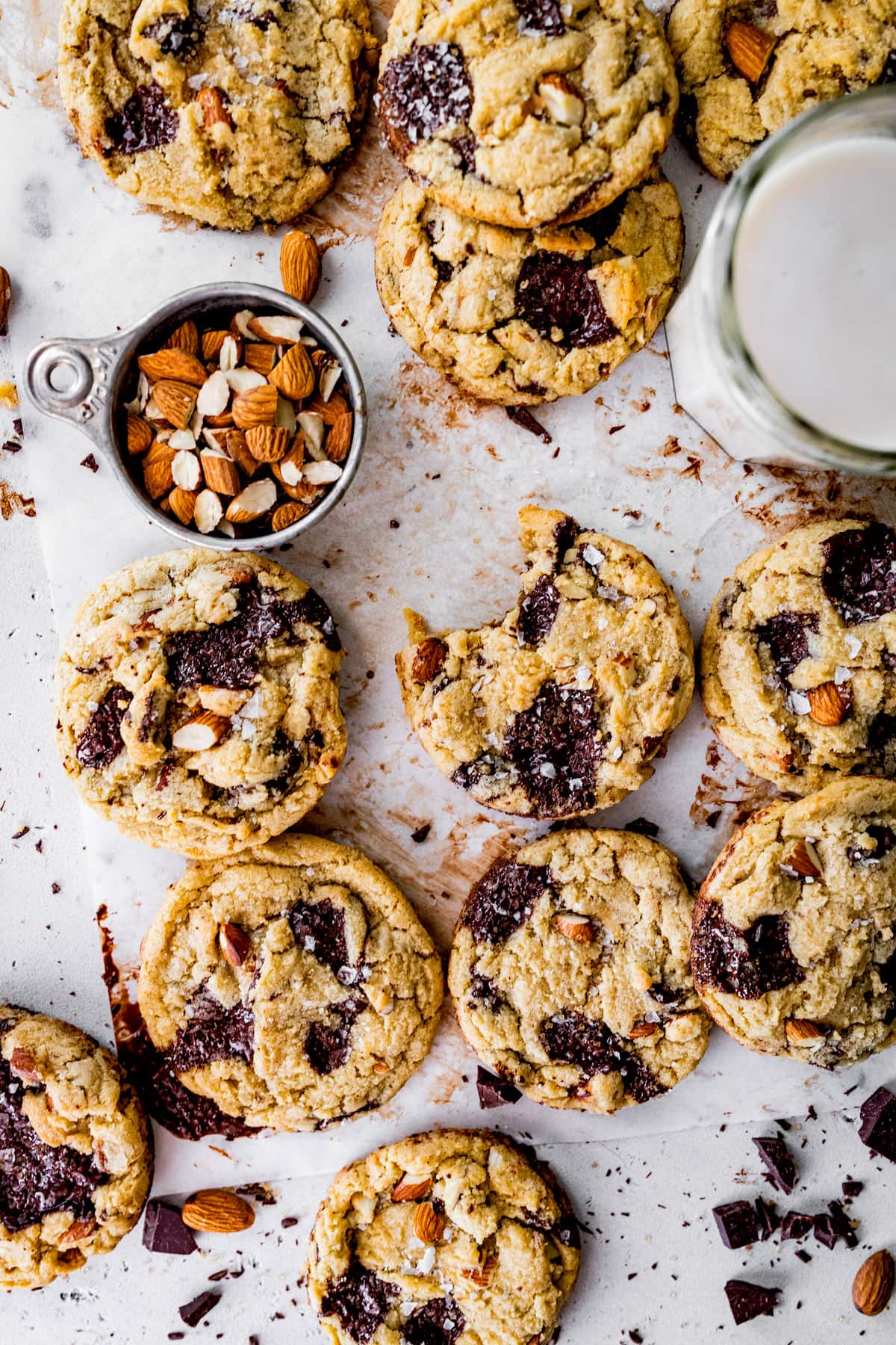 almond chocolate chip cookies on parchment paper with cup of chopped almonds and glass of milk.