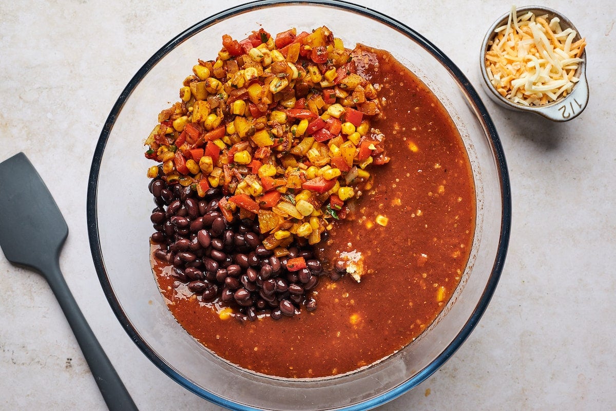 veggies, black beans, enchilada sauce, and quinoa in glass mixing bowl with spatula and cup of cheese on the side. 