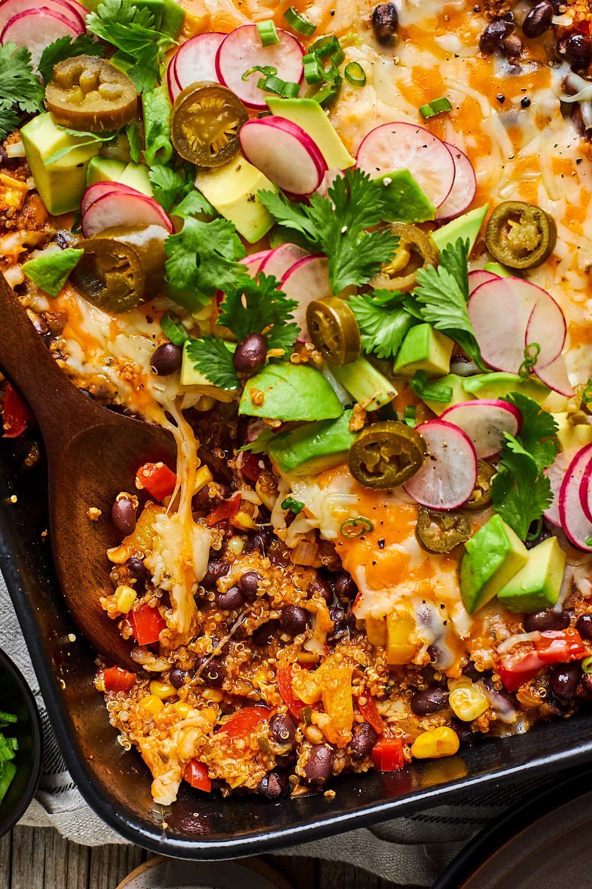 black bean enchilada bake in pan with cheese, toppings, and wooden spoon scooping the casserole.