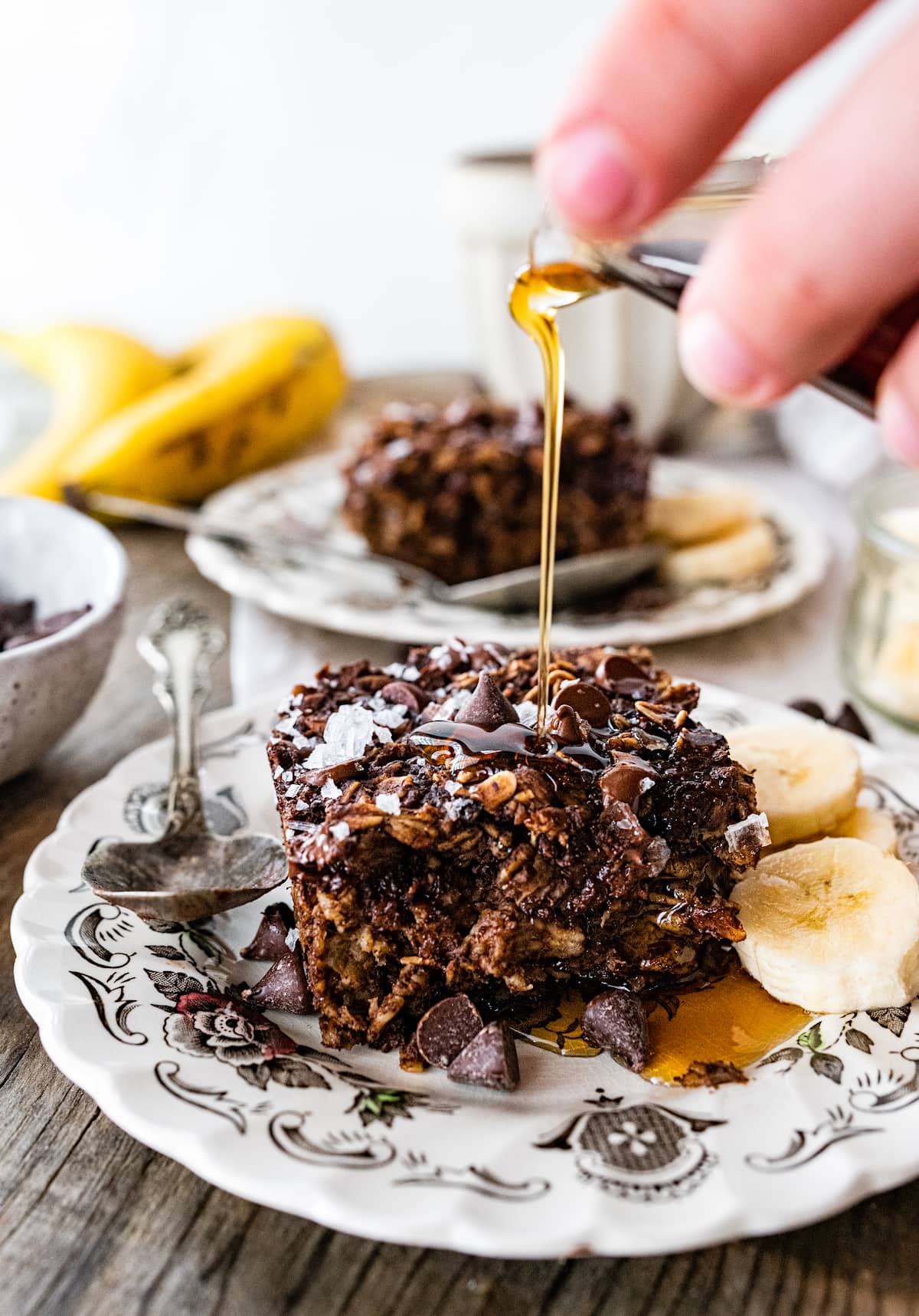chocolate banana baked oatmeal square on plate with spoon, chocolate chips, banana slices, and maple syrup poured on top.