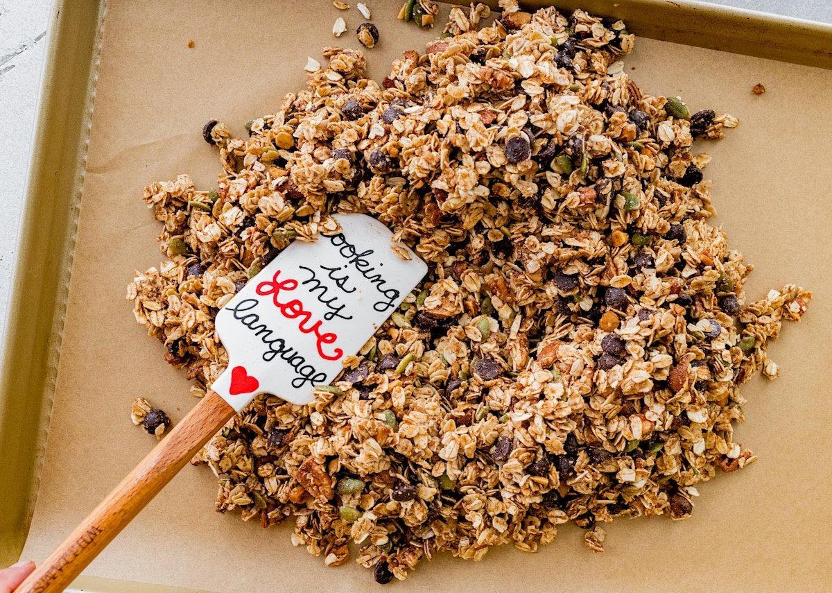 spreading chocolate chip cookie granola on pan with parchment paper with a spatula. 