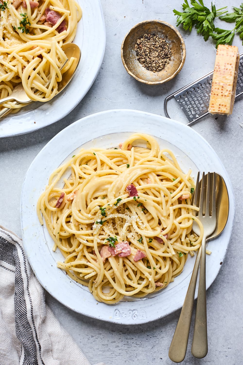spaghetti carbonara on plate with fork and spoon with small bowl of freshly cracked black pepper in bowl and Parmesan cheese.