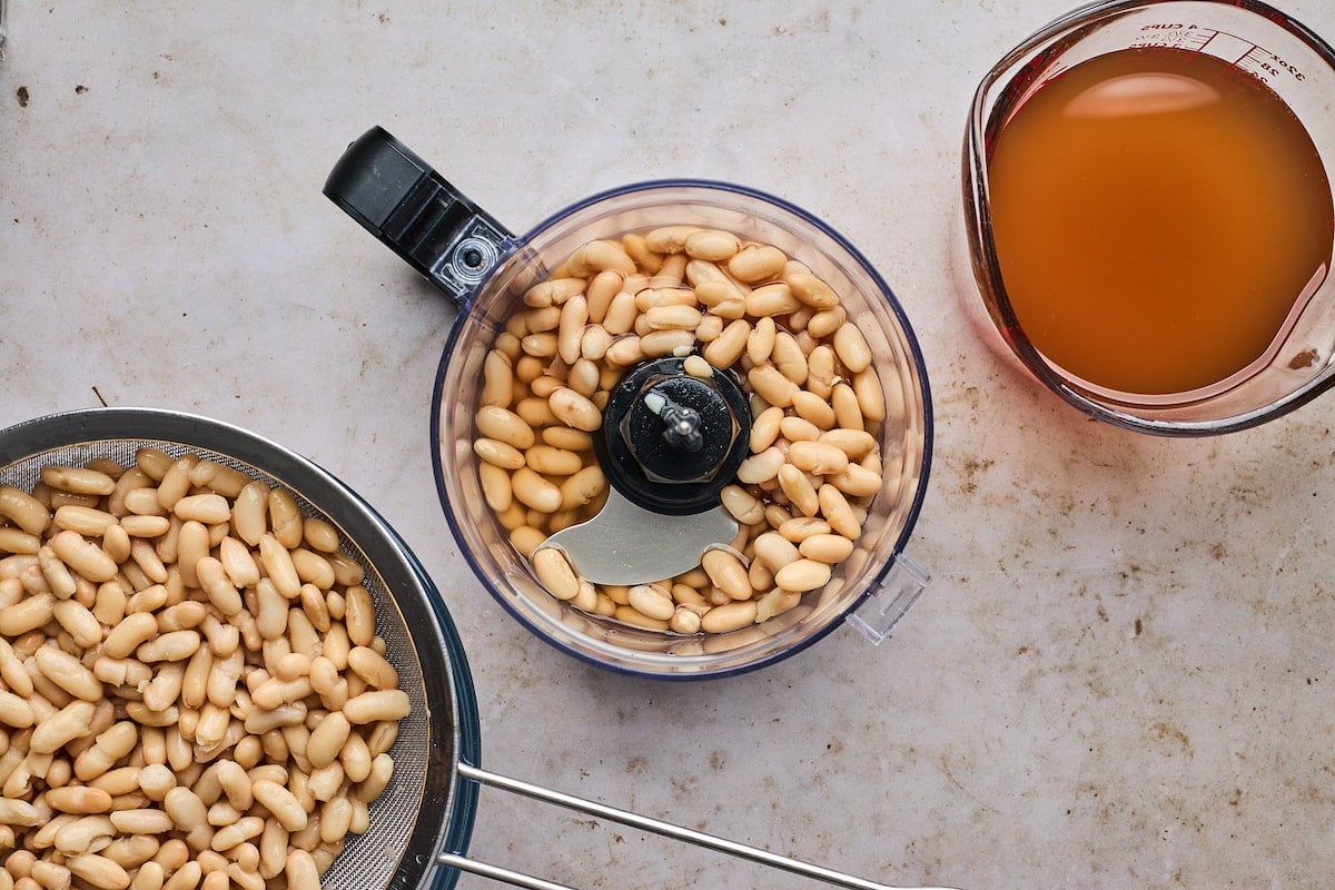 white beans in strainer, white beans in food processor, and vegetable broth in measuring cup.