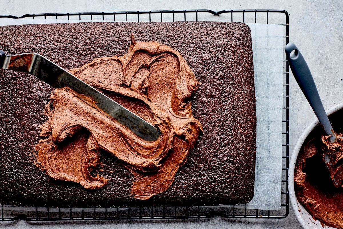 chocolate sheet cake on cooling rack being frosted with chocolate frosting.