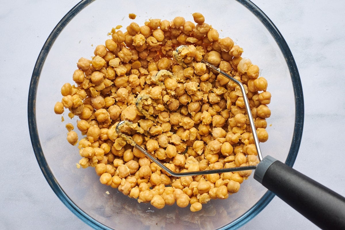 chickpeas being mashed with a potato masher in a large bowl.