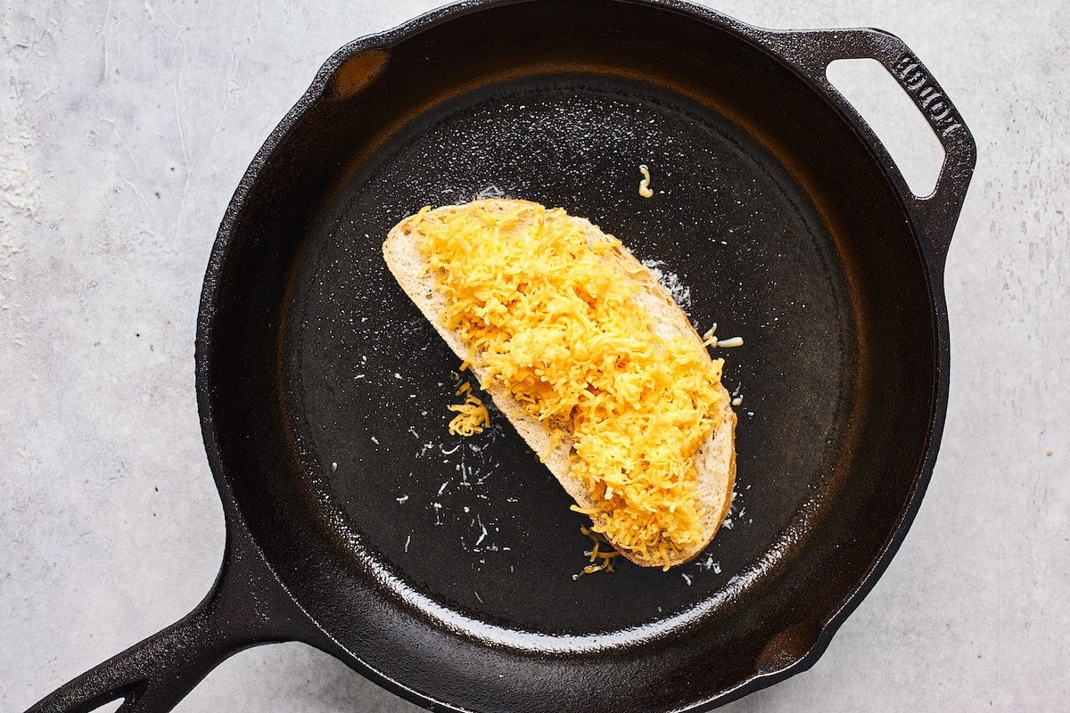piece of bread in cast iron skillet with shredded cheddar cheese to make grilled cheese. 