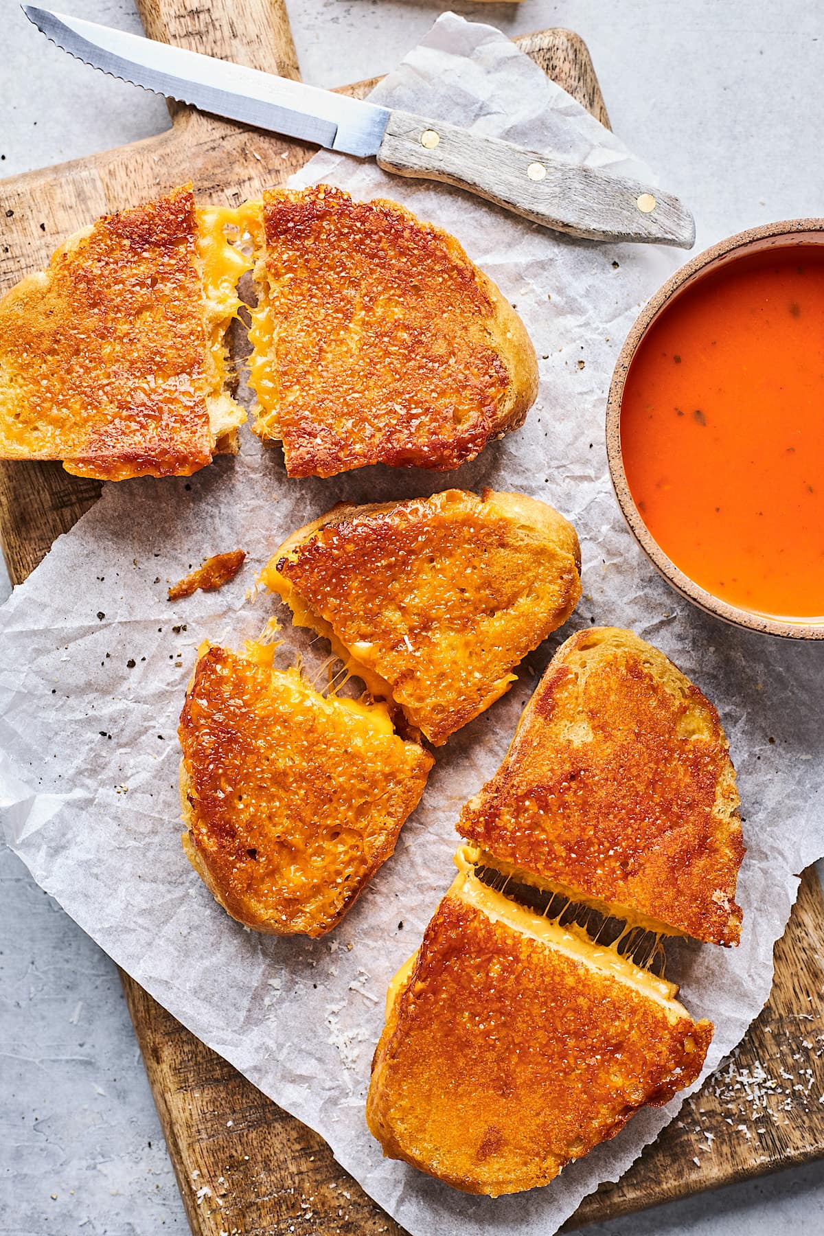 cheese crusted grilled cheese sandwiches on parchment paper on wood board with a bowl of tomato soup.