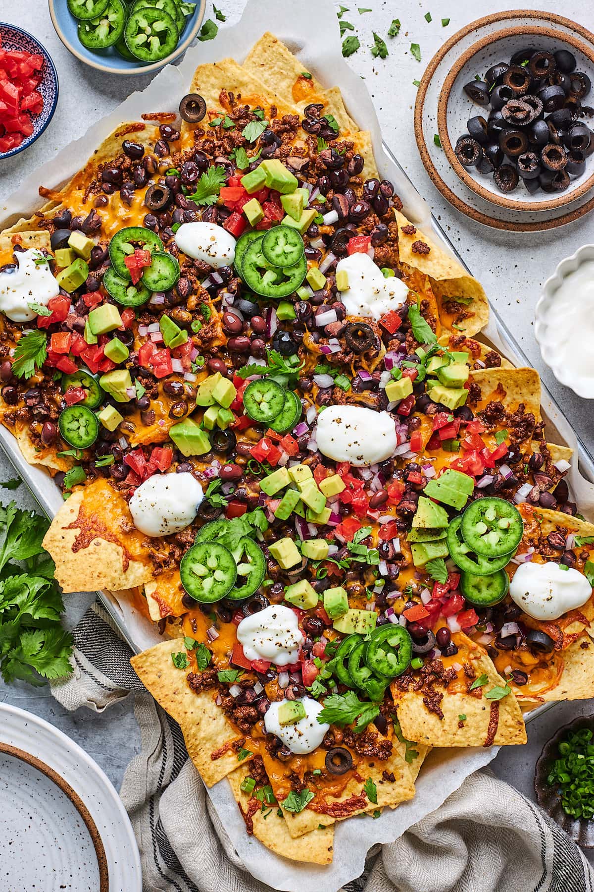 nachos on sheet pan with cheese, ground beef, beans, tomatoes, avocado, sour cream, onion, olives, and jalapeño. 